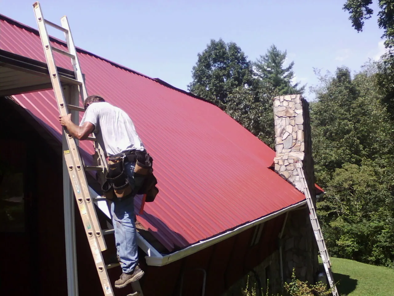 Roofer on a ladder beside a red metal roof and stone chimney — Soffit & Fascia Repair by Roofing Brothers in Christiana